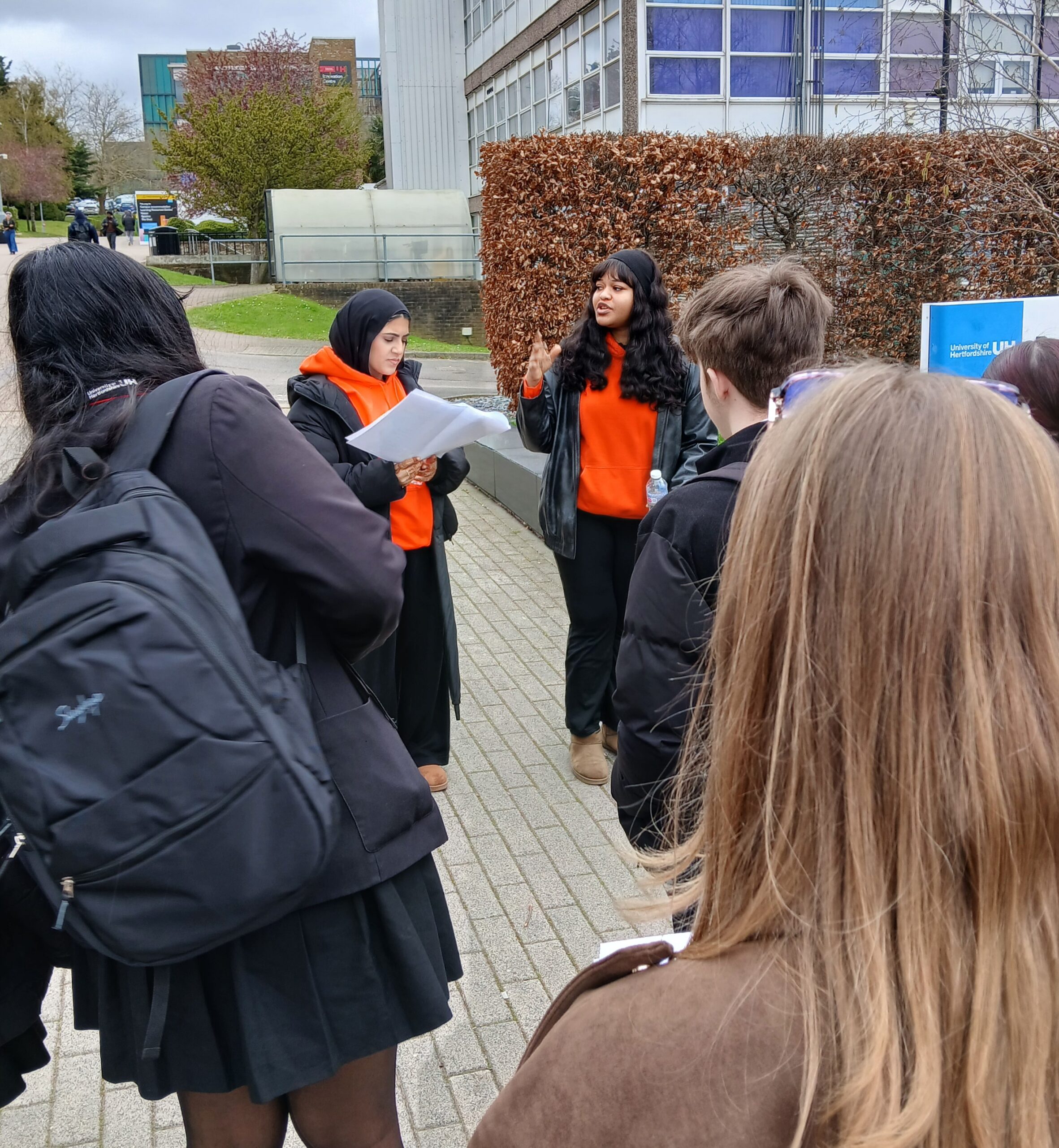 Two university ambassadors in orange WASS hoodies talking to young people on a campus tour. Students listening can be seen from behind